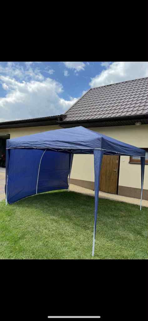 A blue pop-up tent set up on a grassy area near a house with a tiled roof and partly cloudy sky.