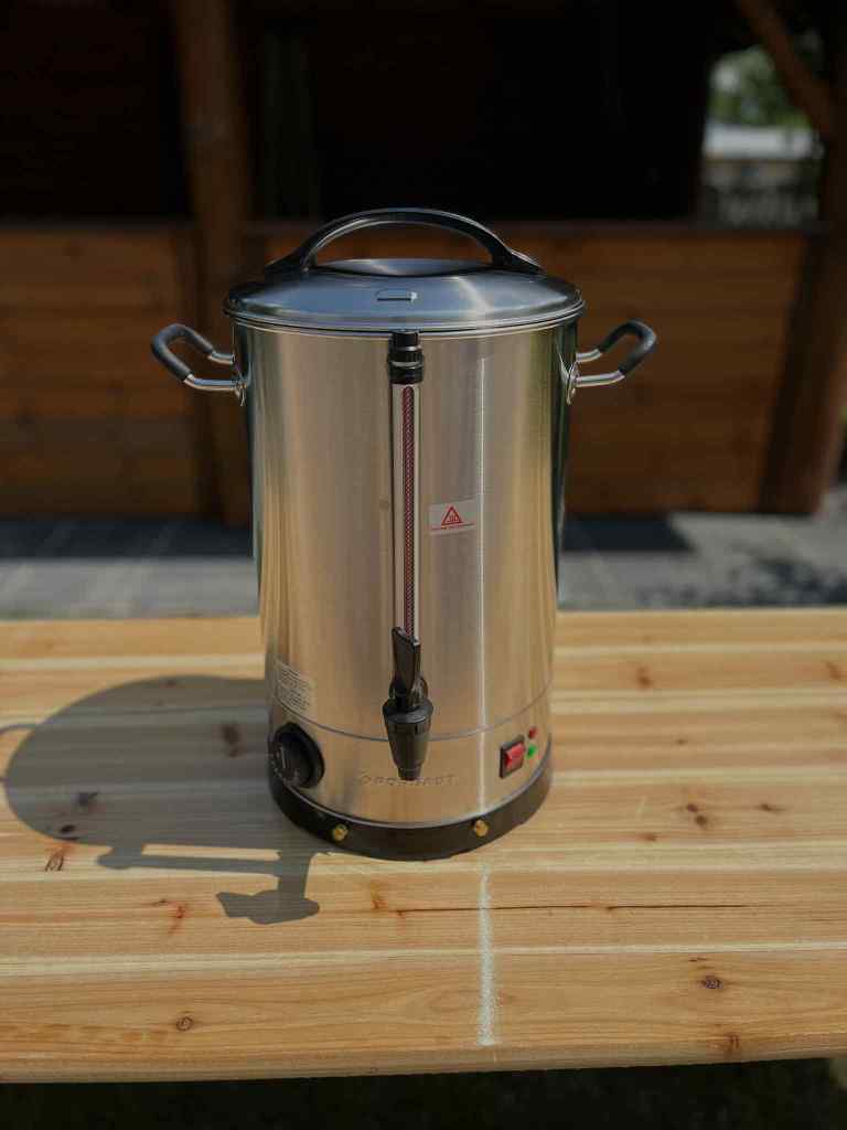 A stainless steel electric kettle with a spigot, placed on a wooden table.