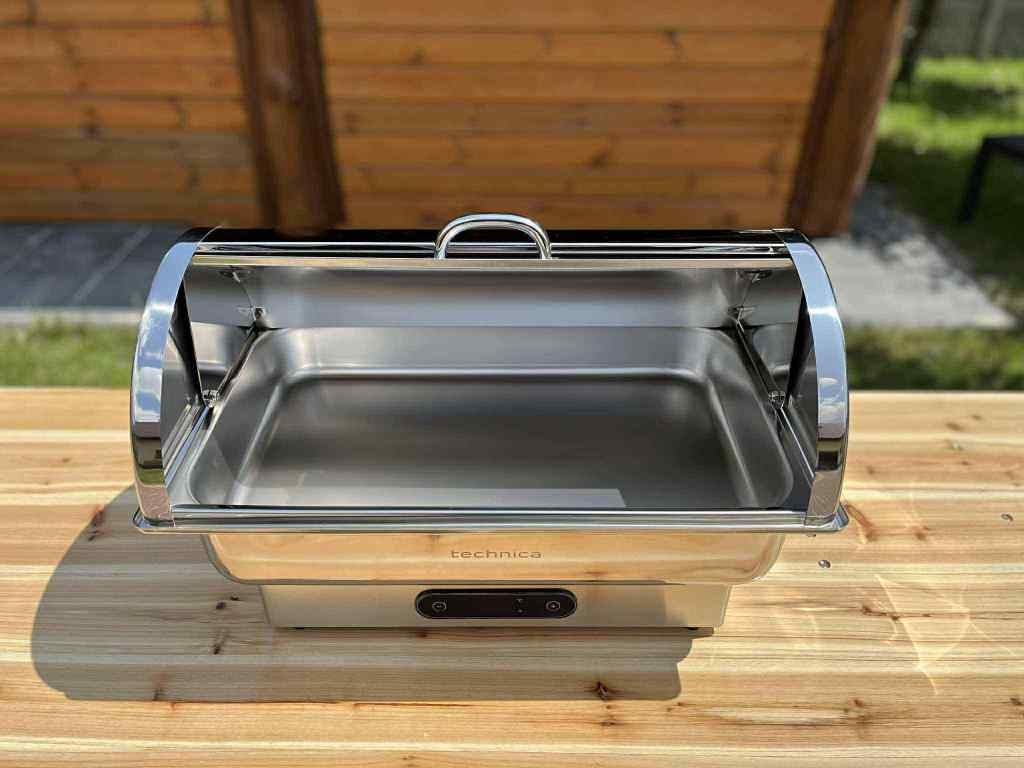 Stainless steel chafing dish with lid open, placed on a wooden table outdoors.