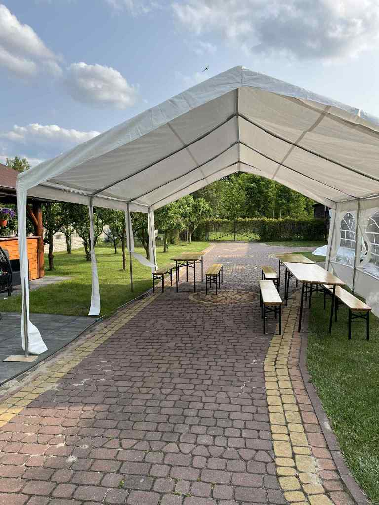 A white tent set up outdoors with wooden tables and benches arranged underneath, on a brick pathway surrounded by greenery and trees.