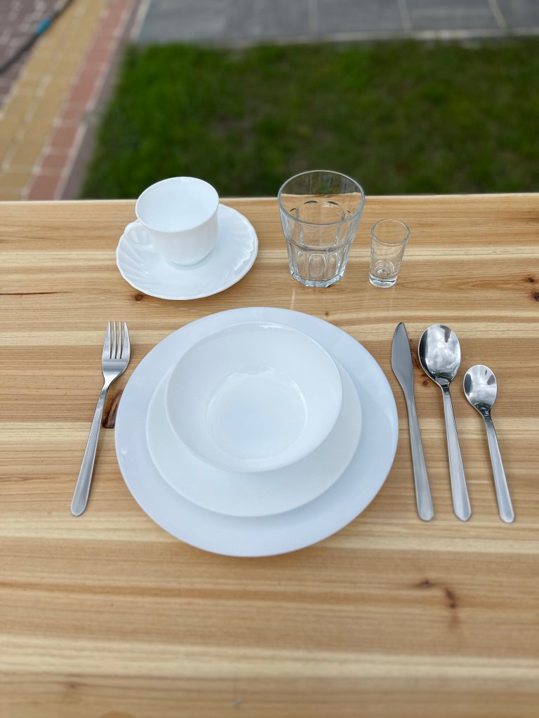 Table setting with white dishes, a cup and saucer, assorted glassware, and cutlery on a wooden surface.