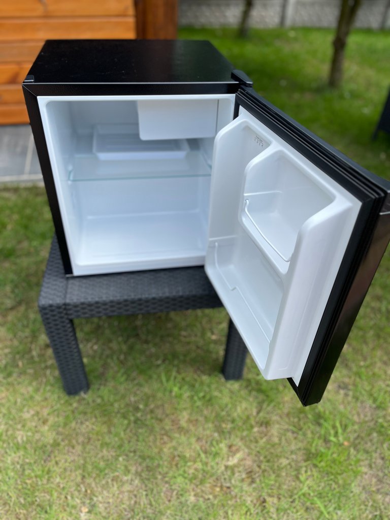 A black mini refrigerator with an open door, revealing a clean, empty interior and a transparent shelf, placed on a wicker table in a grassy outdoor setting.