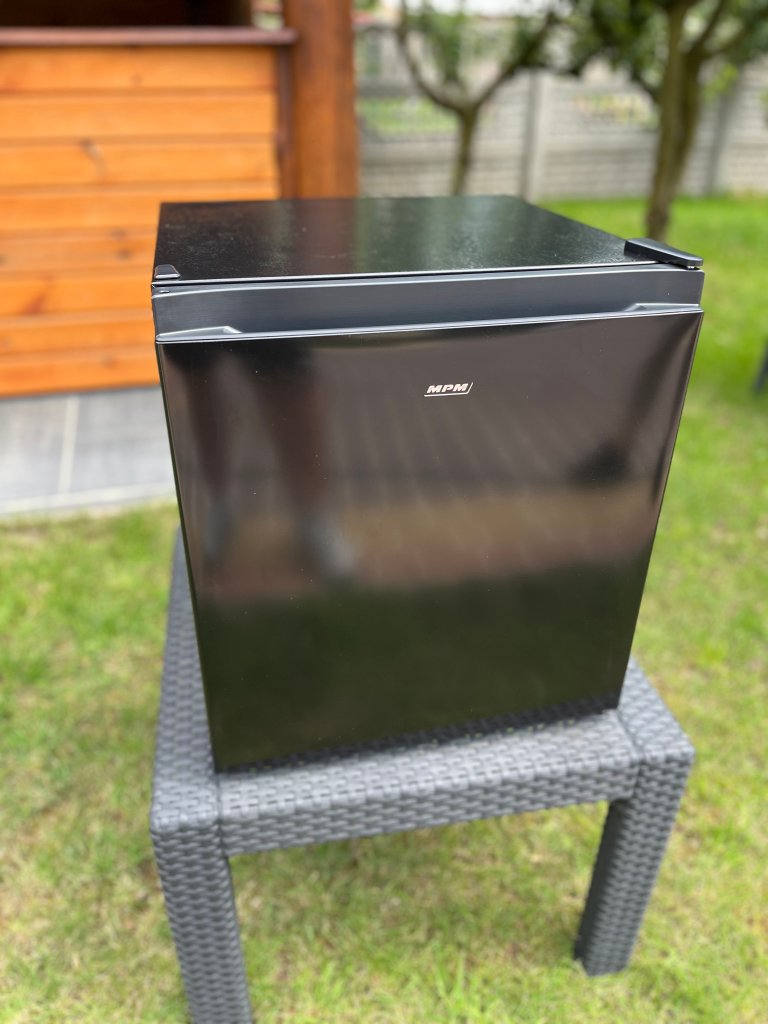 A black mini-fridge placed on a wicker table in a garden setting, with wooden structures and greenery in the background.