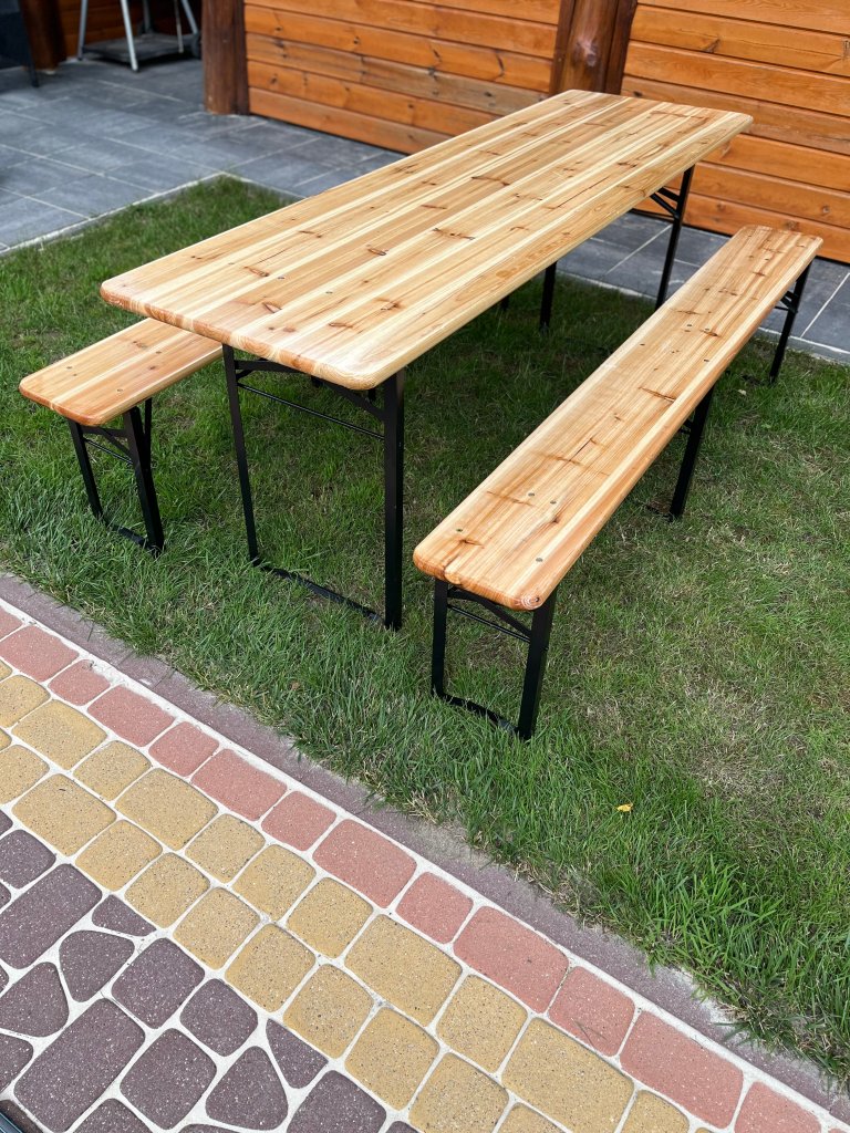 A wooden picnic table with two benches sits on grass next to a paved pathway.