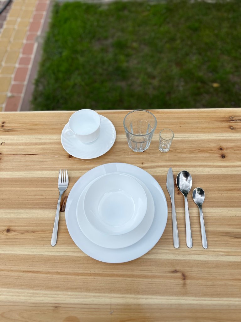 A neatly arranged dining table setting with white plates, a white cup and saucer, a glass, and a small shot glass, all placed on a wooden table with a green lawn in the background.